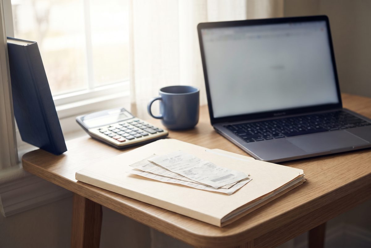 A tidy home-office desk with a laptop, a folder of receipts and a calculator in soft morning daylight.